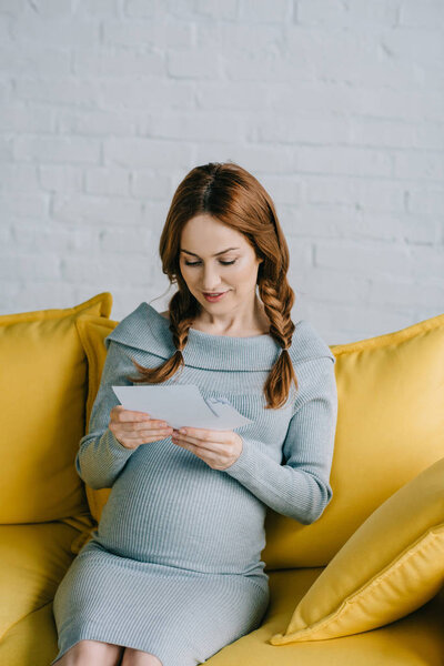 attractive pregnant woman sitting on sofa and looking at photos in living room