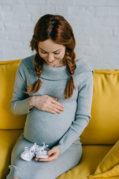 attractive pregnant woman touching abdomen and looking at newborn shoes in living room