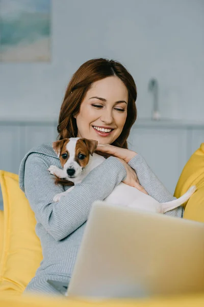 attractive pregnant woman hugging dog and looking at laptop in living room