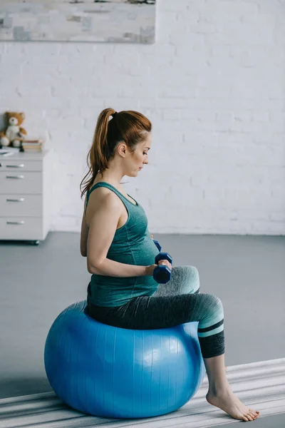 美孕女子在客厅健身球上用哑铃锻炼的侧面观察 — 图库照片