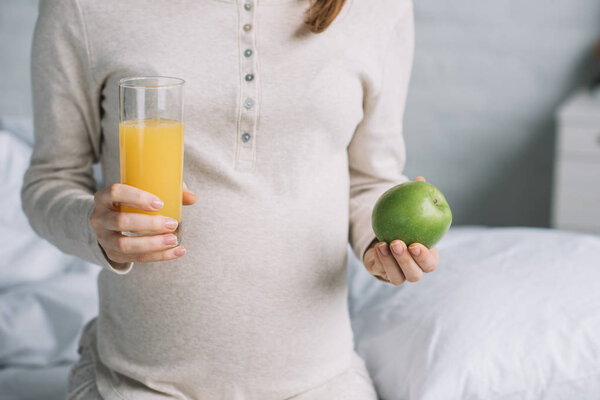 cropped image of pregnant woman holding glass of orange juice and apple in bedroom 