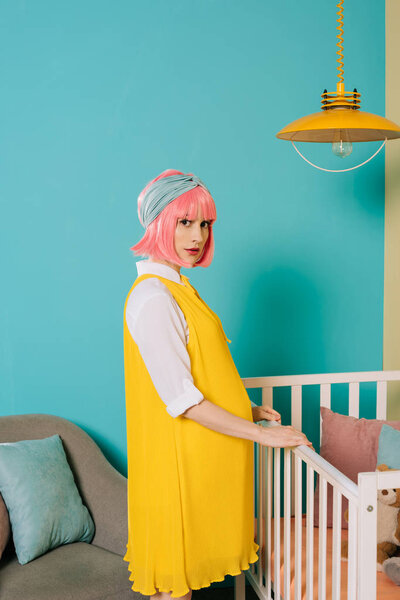 stylish pregnant pin up woman with pink hair standing near baby cot in child room