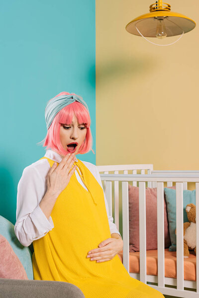 shocked retro styled pregnant woman with pink hair sitting near baby cot in child room