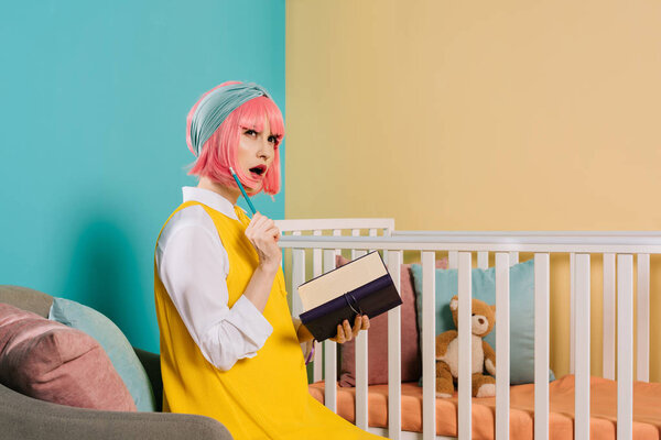 surprised stylish pregnant pin up woman with pink hair holding notebook and pencil near baby cot