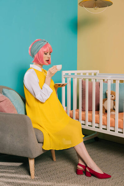 retro styled pregnant pin up woman with pink hair drinking tea near baby cot in child room