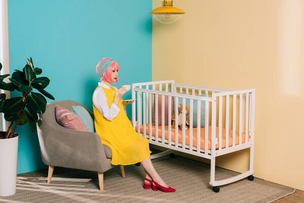 side view of retro styled pregnant pin up woman sitting near baby cot with cup of tea in child room