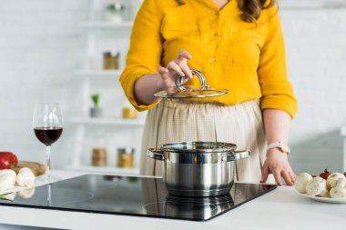 cropped image of woman holding lid from pan near electric stove in kitchen clipart