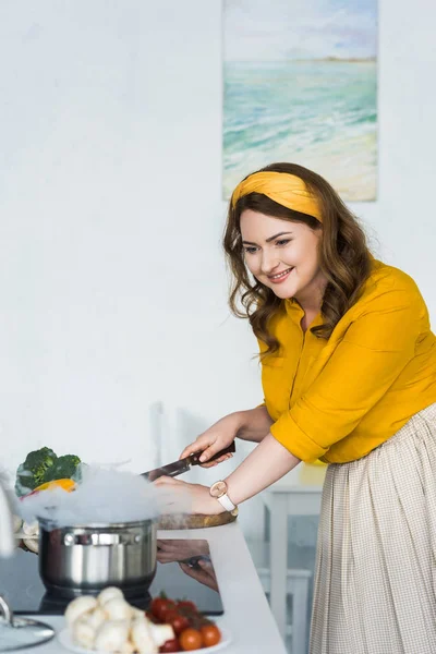 美丽的妇女切割蔬菜和看锅在厨房电炉 — 免费的图库照片