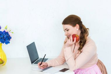 beautiful woman writing something to notebook and holding apple at kitchen clipart