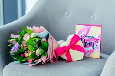 close up view of wrapped bouquet of flowers, heart shaped gift and i love you mom postcard on armchair, mothers day concept