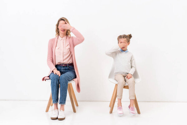 mother and daughter sitting on chairs and covering eyes with hands on white