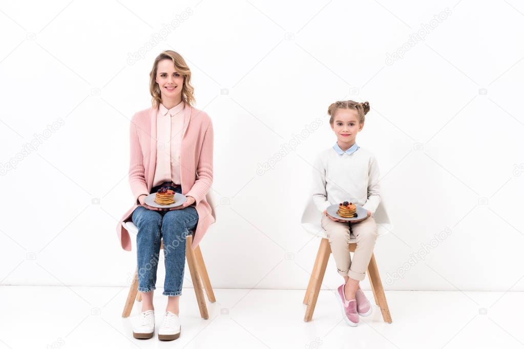 mother and daughter sitting on chairs and holding plates with pancakes on white
