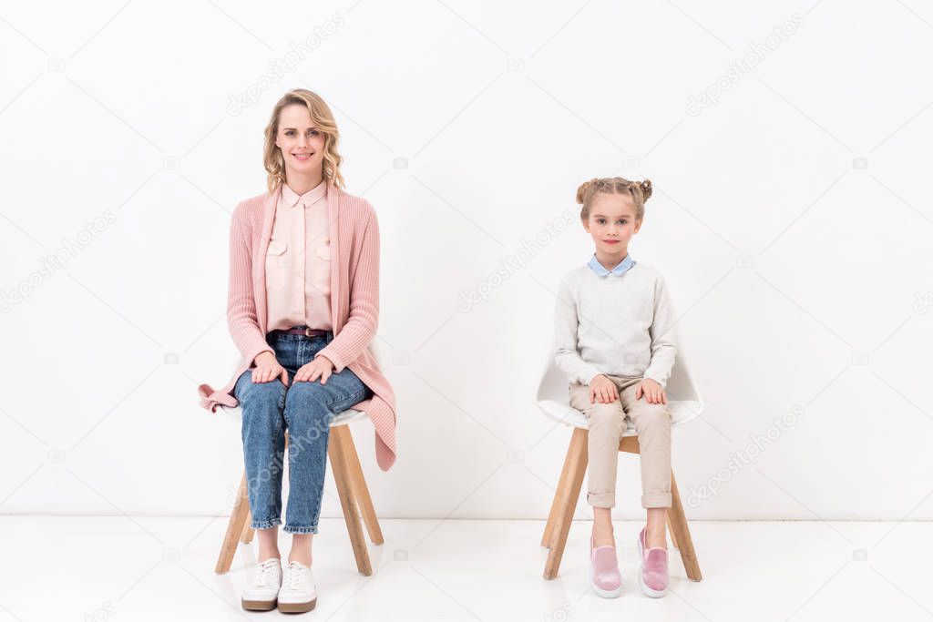 mother and daughter sitting on chairs and looking at camera on white