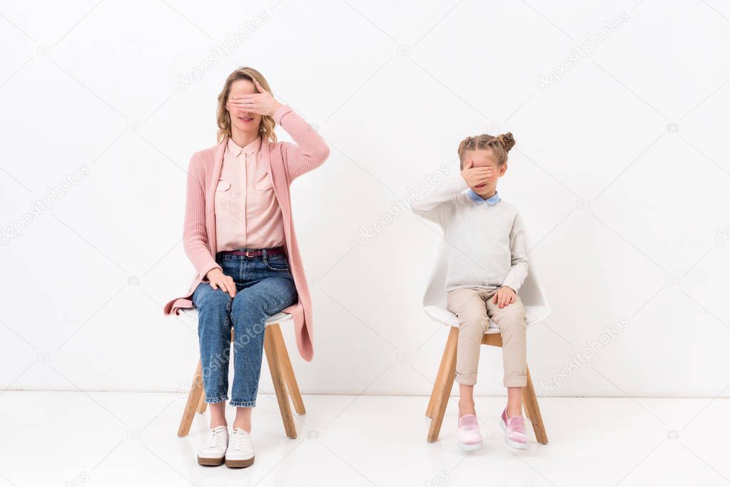 mother and daughter sitting on chairs and covering eyes with hands on white