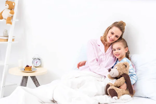 mother and daughter lying in bed with teddy bear at home