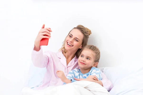 微笑的母亲和女儿带自拍智能手机在家里的床上 — 图库照片