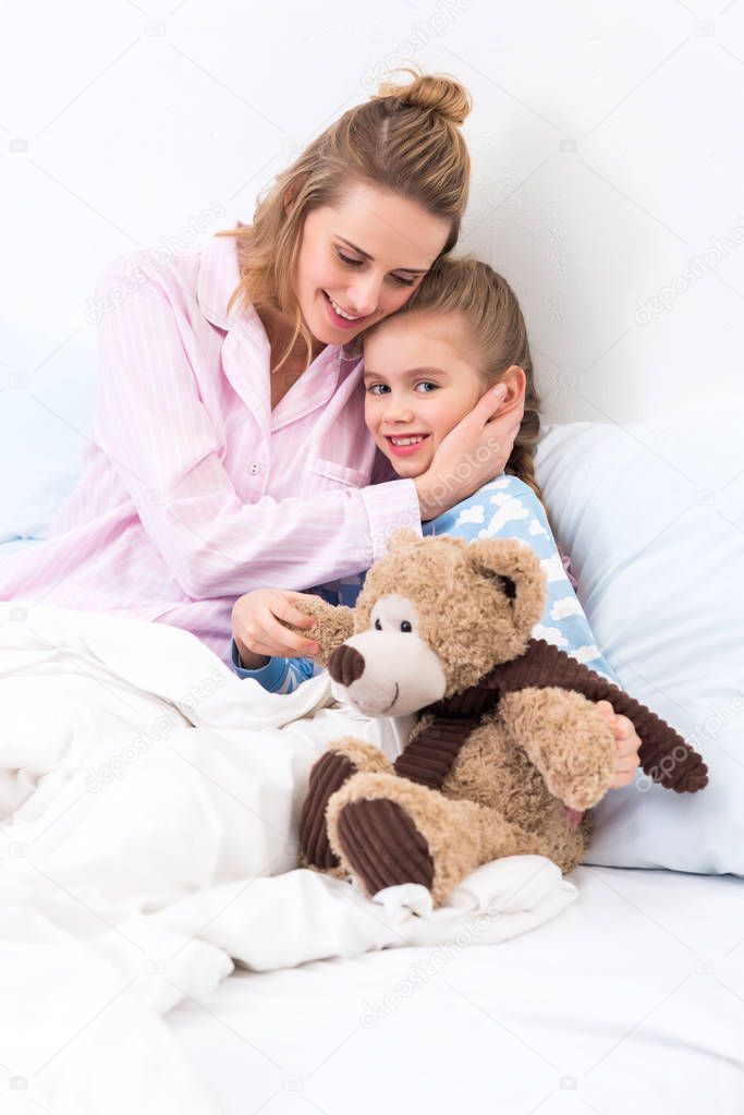 mother hugging daughter with teddy bear on bed at home