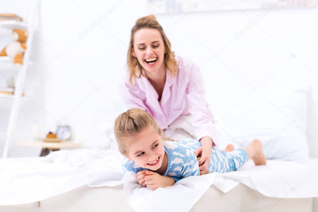 smiling mother tickling daughter on bed at home