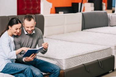 smiling couple with notebook sitting on mattress in furniture store clipart