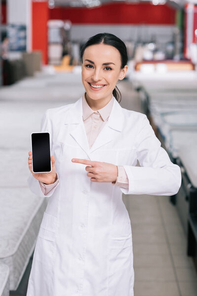 smiling shop assistant in white coat showing smartphone with blank screen in hand in furniture shop with mattresses