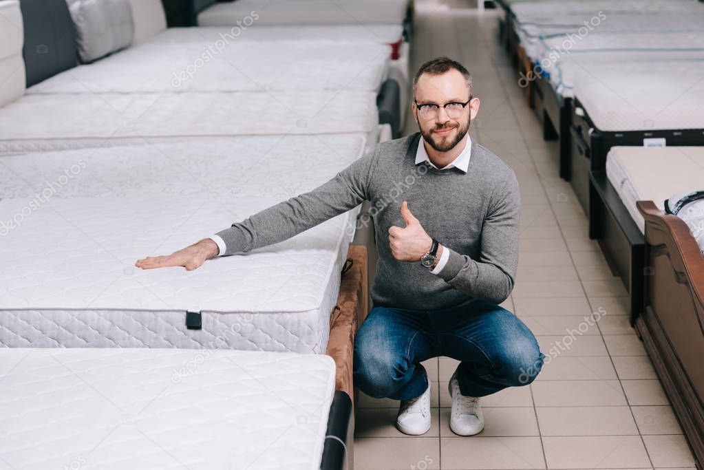 male customer in eyeglasses showing thumb up while touching orthopedic mattress in furniture shop