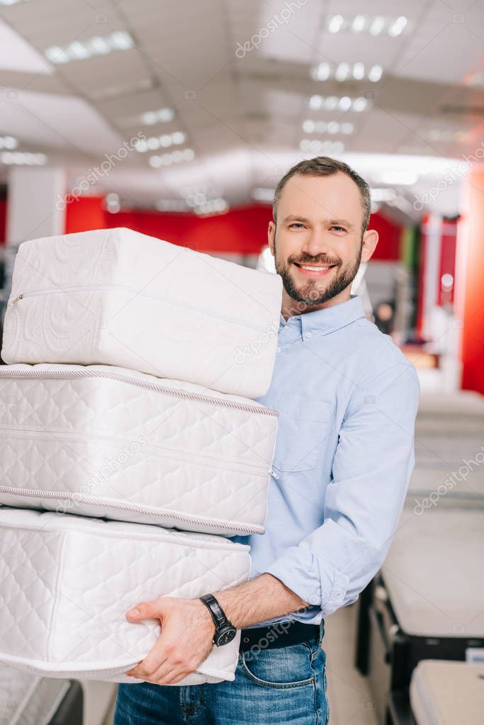 smiling man holding pile of folding mattresses in hands in furniture store 