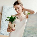 Schöne junge Künstlerin lehnt an Bild auf Staffelei und betrachtet grüne Topfpflanze