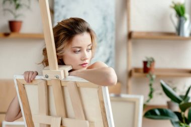 beautiful sensual young woman leaning at easel and looking away in art studio clipart