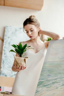 beautiful young artist leaning at picture on easel and looking at green potted plant clipart