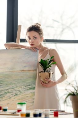 Saksılı bitki tutarak ve şövale art Studio, yaslanmış güzel genç kadın ressam