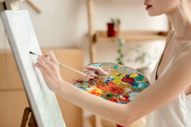 cropped shot of tender young artist with palette and brush painting on easel in studio clipart