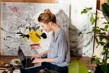 side view of smiling young female painter using laptop in art studio clipart