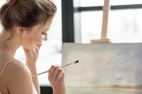 красивая улыбающаяся молодая женщина держит кисть и смотрит вниз в художественной студии
