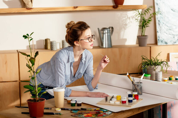 young female artist in eyeglasses standing at table with empty canvas and painting supplies 
