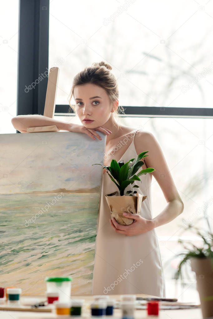 beautiful young female painter holding potted plant and leaning at easel in art studio
