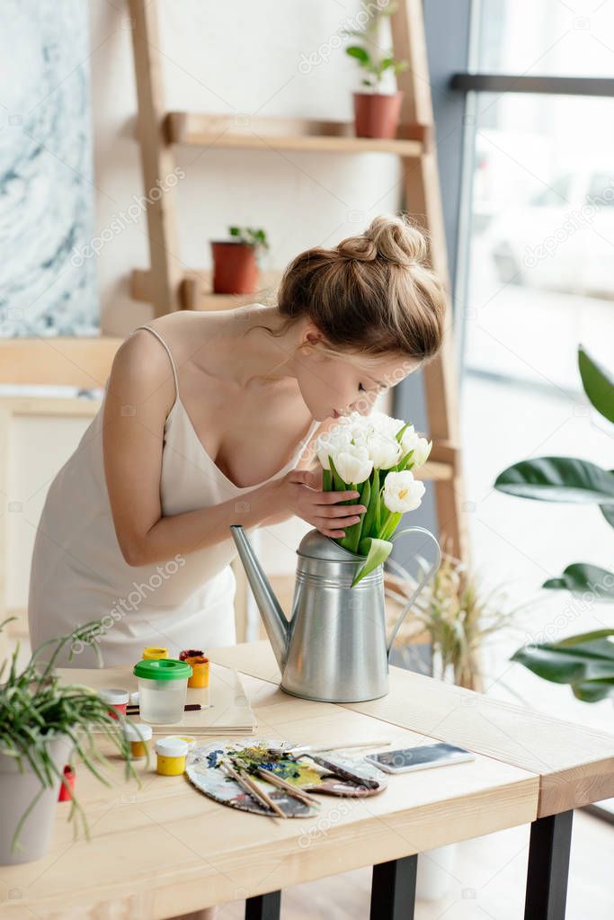 beautiful young woman sniffing flowers in watering can in art studio