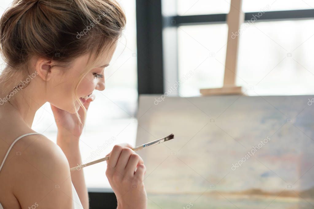beautiful smiling young woman holding brush and looking down in art studio