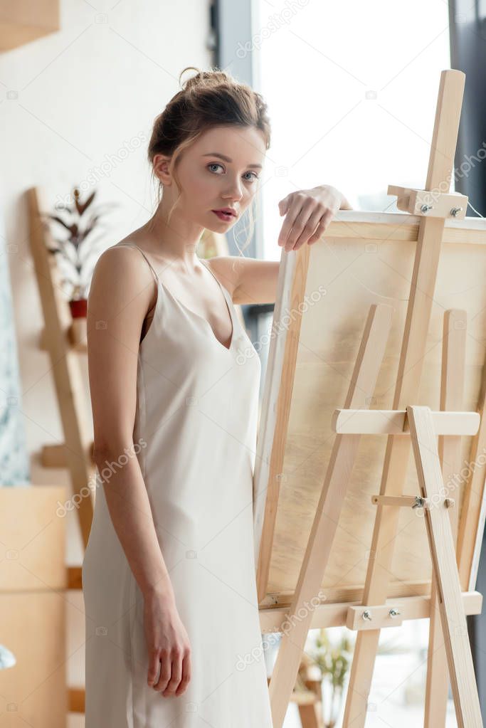 beautiful tender young artist leaning at easel and looking at camera