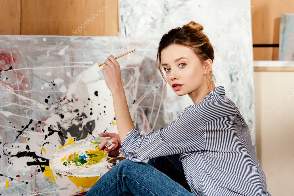 beautiful young artist holding palette and looking at camera in art studio