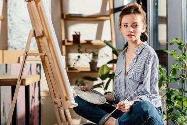 attractive female artist sitting on bench and holding palette and paintbrush clipart