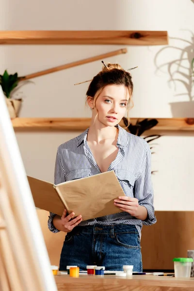 Atractiva Artista Femenina Leyendo Libro Pie Mesa Con Suministros Pintura — Foto de stock gratis