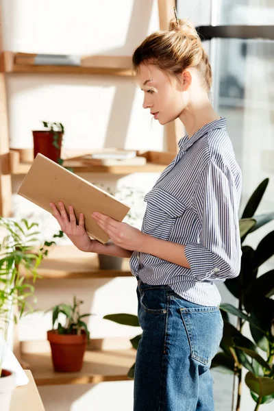 若い女性は 鉢植えの植物の近くの本を読んでの側面図 — ストック写真