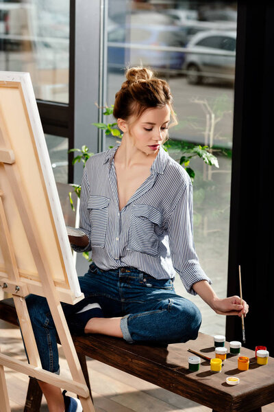 женщина-художник сидит на скамейке с красками и рисует картину на мольберте
 