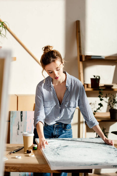 female artist looking at own painting on table with coffee cup 
