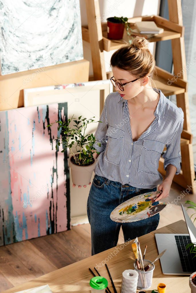 stylish female artist in eyeglasses holding plant and palette in studio 