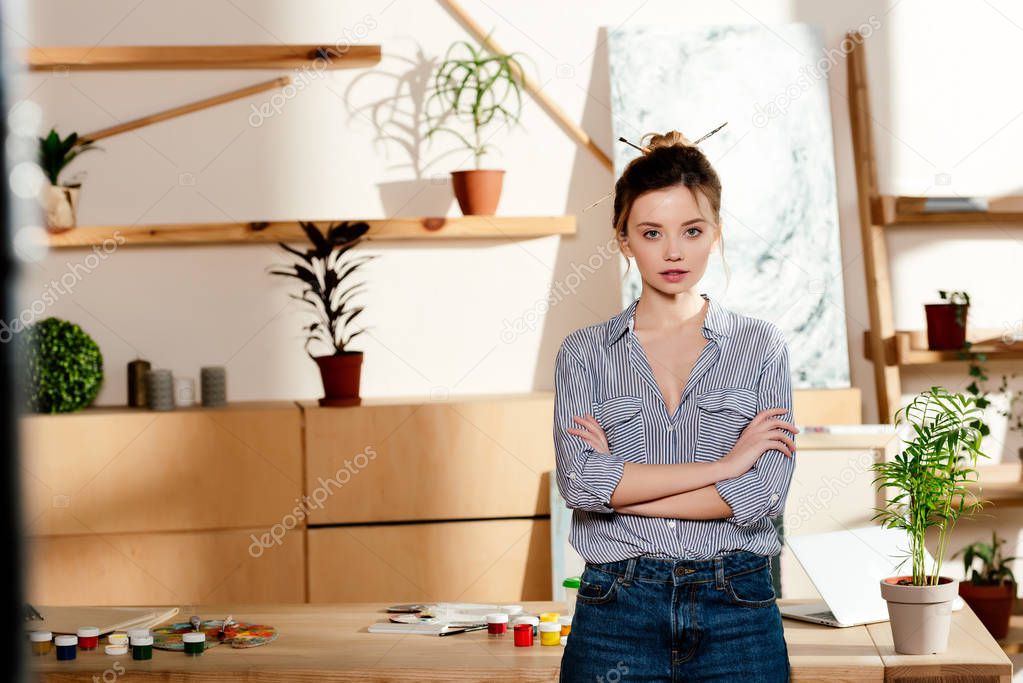 portrait of young attractive female artist with crossed arms standing near table with painting supplies 