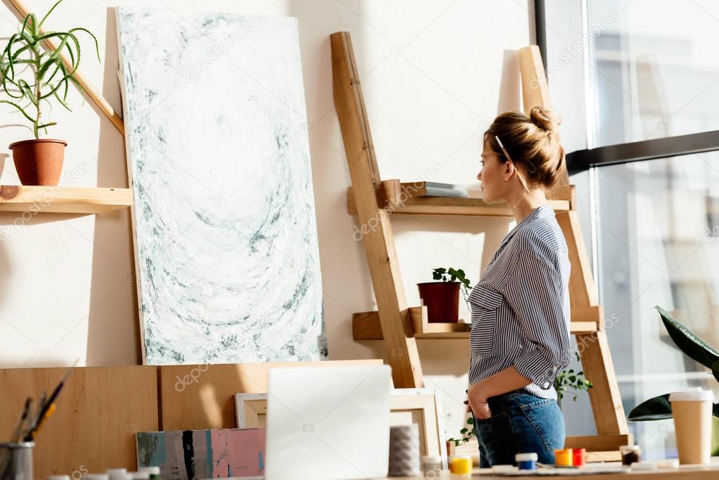 female artist with paintbrush behind ear looking at own picture in studio 
