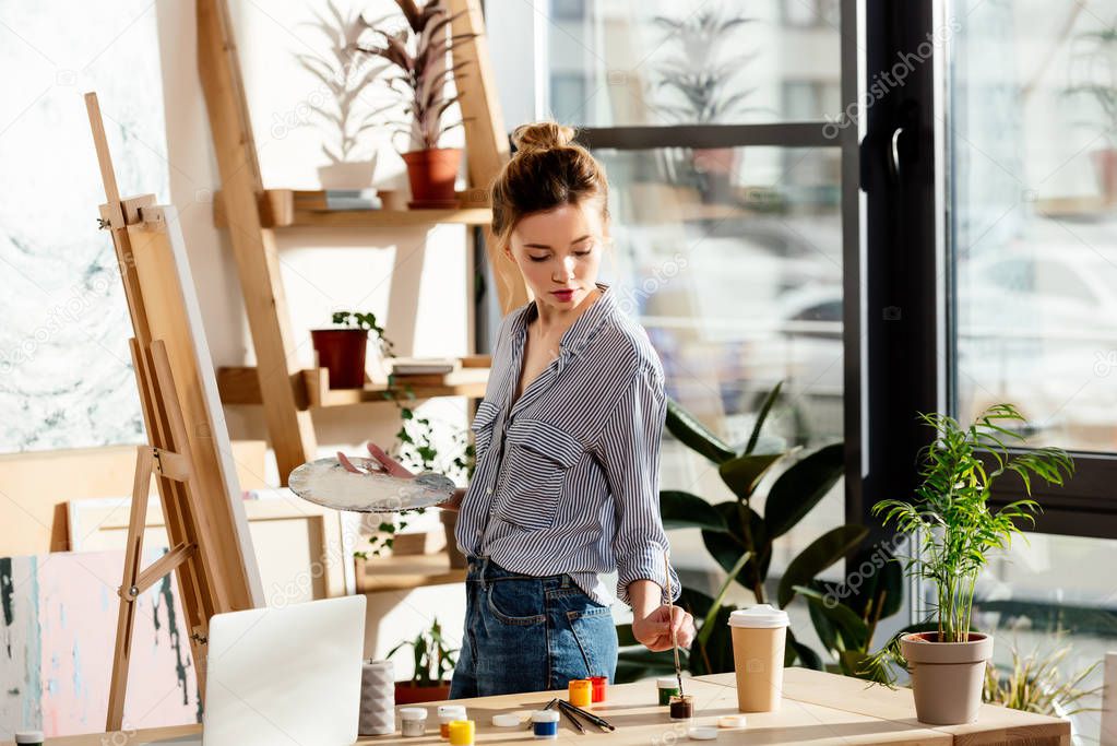 female artist holding palette and putting brush into paint in studio