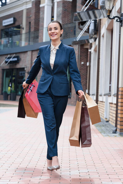smiling attractive businesswoman walking with paper shopping bags on street