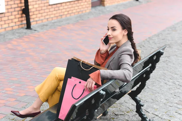 side view of beautiful woman sitting on bench with shopping bags and talking by smartphone